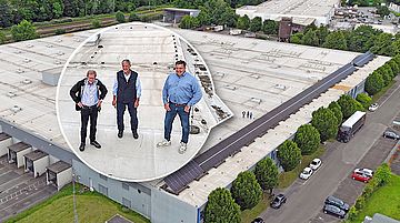 Drei Männer stehen auf dem Dach eines großen Industriegebäudes mit Solarpaneelen, umgeben von Bäumen und Parkplätzen mit Fahrzeugen